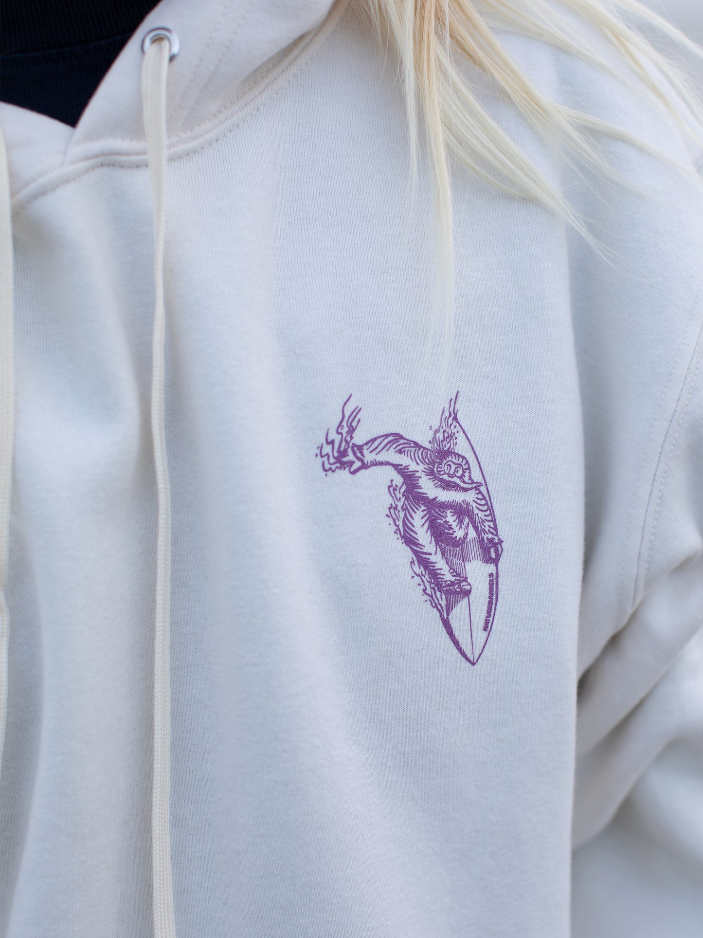Close-up of a white hoodie with a small purple chest graphic of a surfing sasquatch