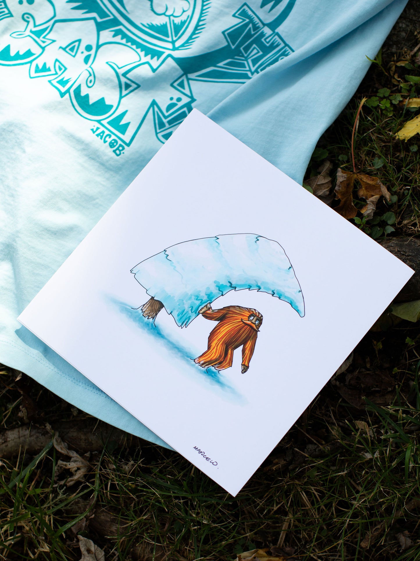 A vibrantly colored illustration from 'The Book of Barrels' showing an orange sasquatch under a snow-covered pine tree placed atop the back of a light blue t-shirt with a large dark blue graphic. The t-shirt and book lie on the natural forest floor.