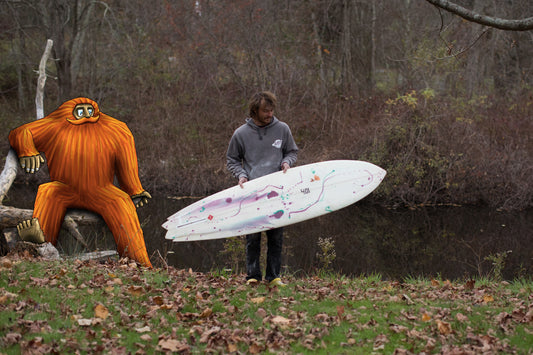 A person holding a surfboard next to a large illustrated orange sasquatch, set against a woodland backdrop