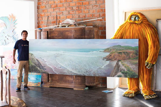 A man standing beside a big orange sasquatch character, both holding the ends of a large painting of a coastal surf scene in an art studio.