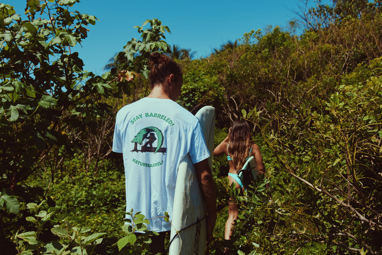 A person walking through a lush green path carrying a surfboard wearing a white t-shirts that reads 'STAY BARRELED! NATUREBARRELS' on the back. A woman can be seen carrying a surfboard head of him.