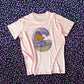 A light pink t-shirt with a large graphic on the back of an orange sasquatch riding a wooden surfboard on a purple wave made of acai liquid coming out of a bowl. The shirt is displayed against a background of acai berries. 