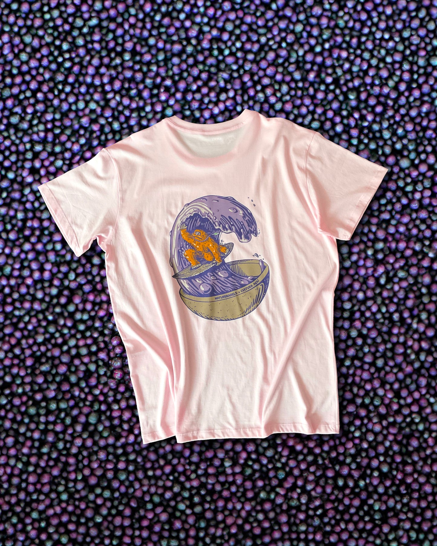 A light pink t-shirt with a large graphic on the back of an orange sasquatch riding a wooden surfboard on a purple wave made of acai liquid coming out of a bowl. The shirt is displayed against a background of acai berries. 