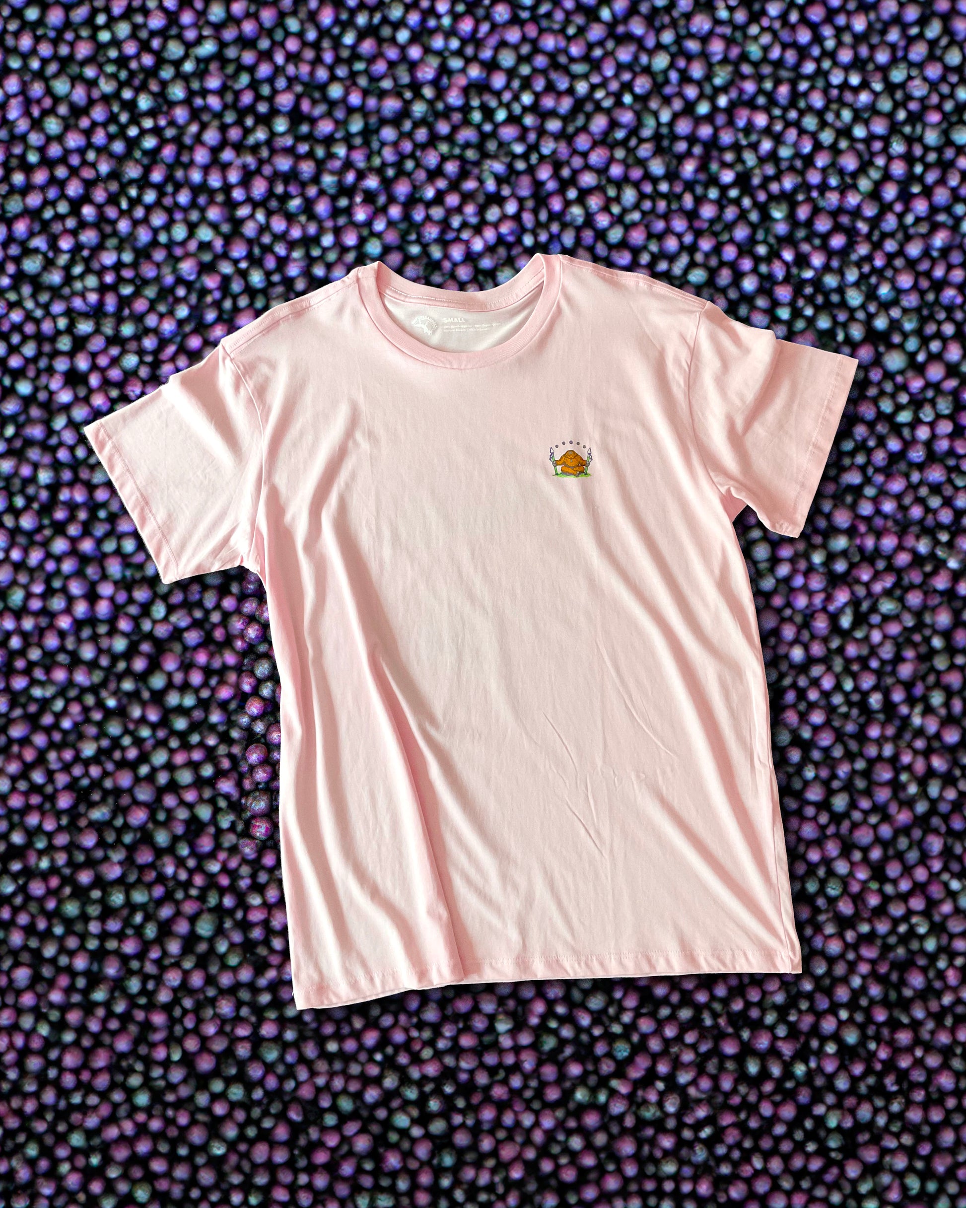A light pink t-shirt with a small chest print of an orange sasquatch sitting on grass, holding two spheres with acai berries hovering above the sasquatch. The t-shirt is lying flat against a background of acai berries. 