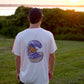 A person wearing a cap and a light pink t-shirt with a back graphic of an orange sasquatch riding a wooden surfboard on a purple wave made of acai liquid coming out of a bowl. The person is standing in a grass field at sunset