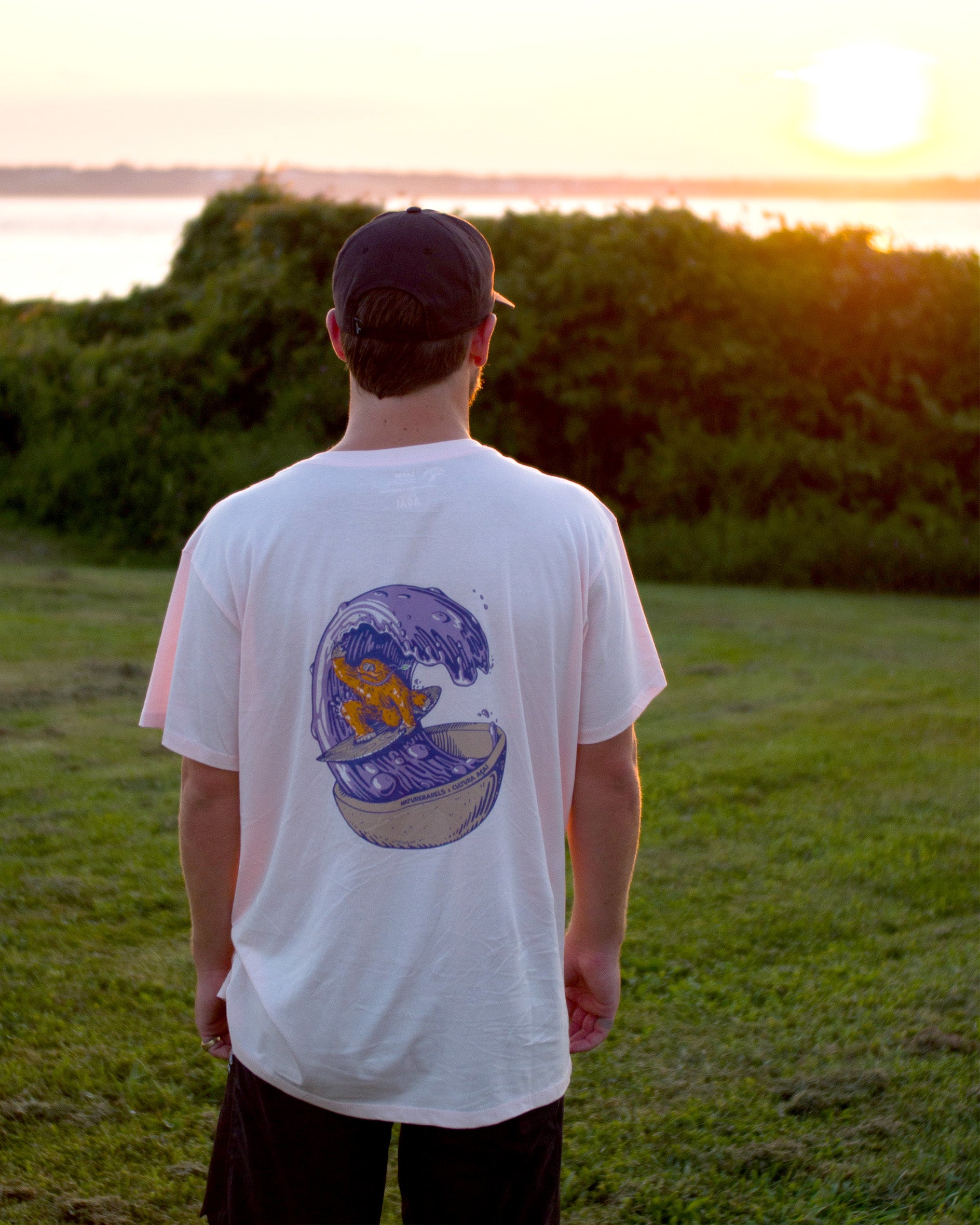 A person wearing a cap and a light pink t-shirt with a back graphic of an orange sasquatch riding a wooden surfboard on a purple wave made of acai liquid coming out of a bowl. The person is standing in a grass field at sunset