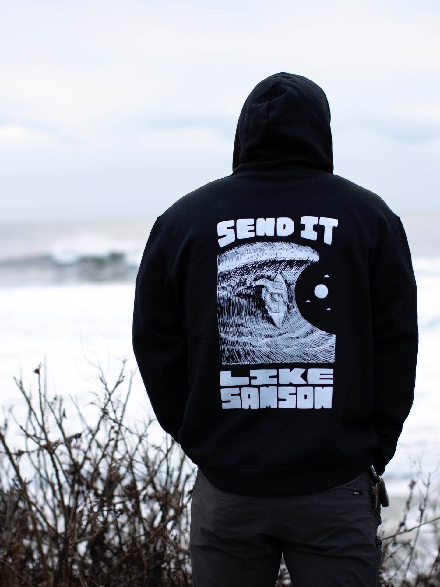 Person standing with their back to the camera lloking at the ocean wearing a black hoodie featuring a large graphic of a surfing sasquatch riding a wave and 'SEND IT LIKE SAMSON' text.