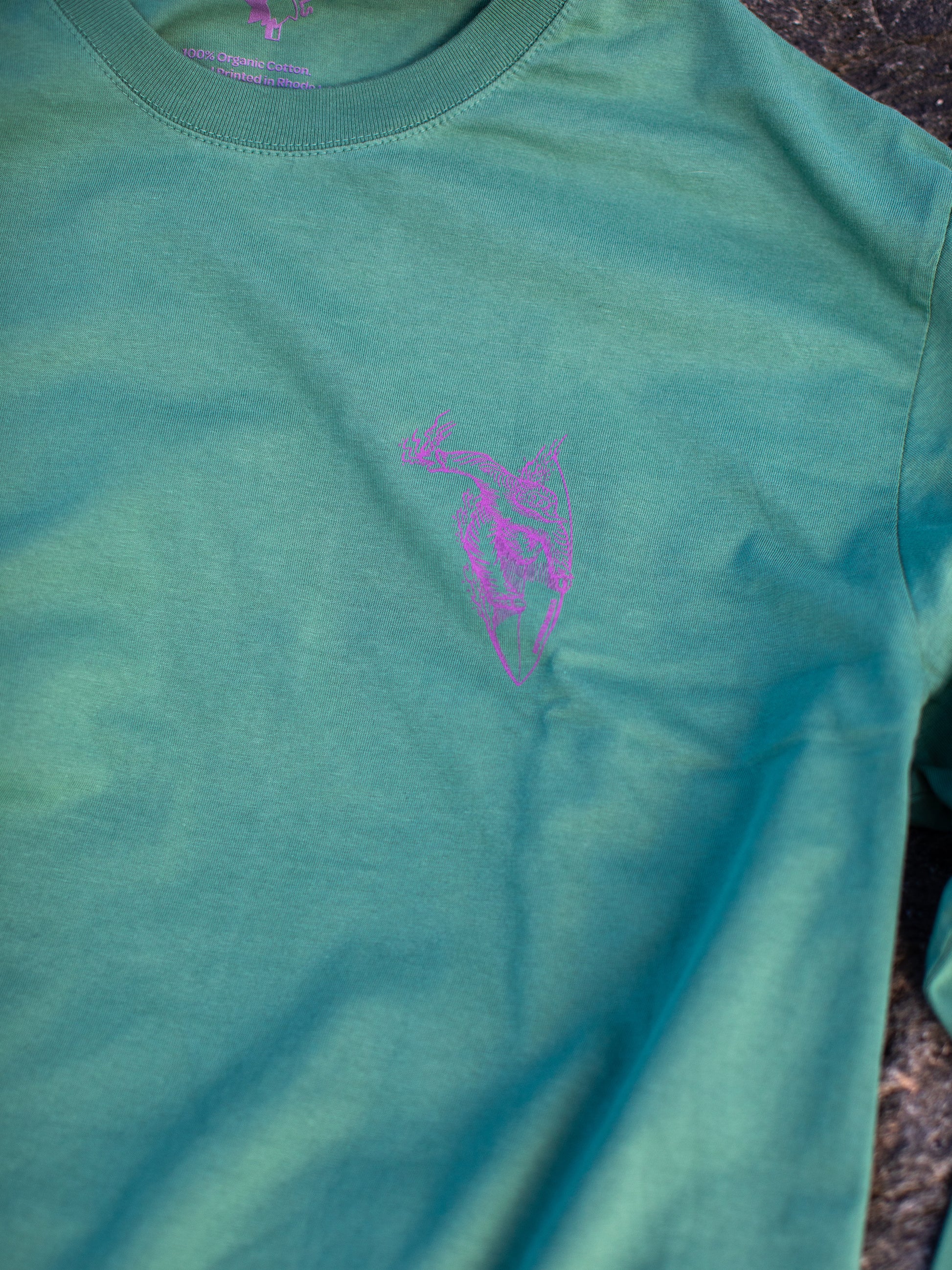 Close-up of a green long sleeve t-shirt with a small purple chest graphic of a surfing sasquatch