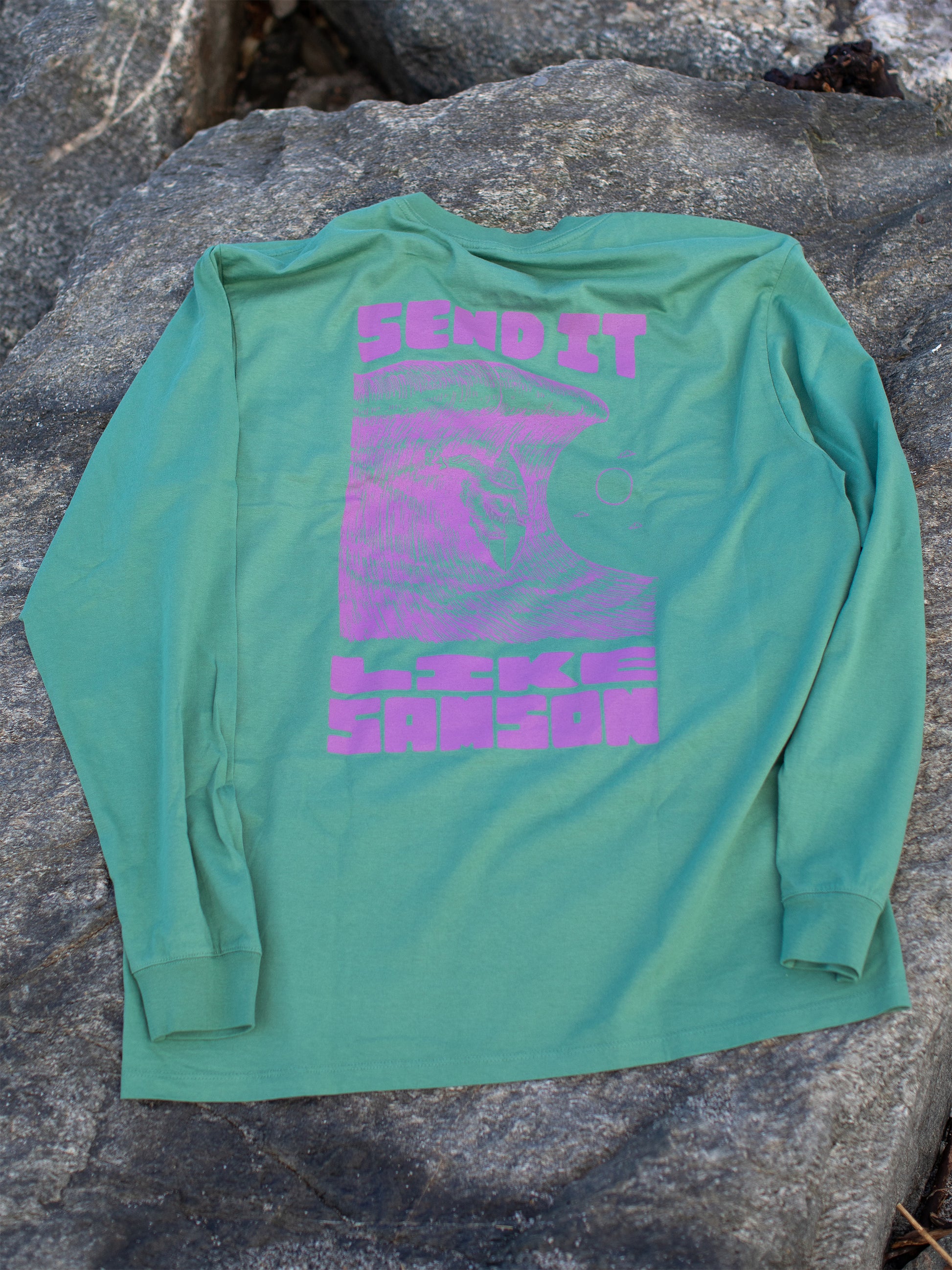 Green long sleeve t-shirt with large purple graphic in the back featuring 'SEND IT LIKE SAMSON' text and an illustration of a sasquatch riding a large wave, laid on a rocky surface.