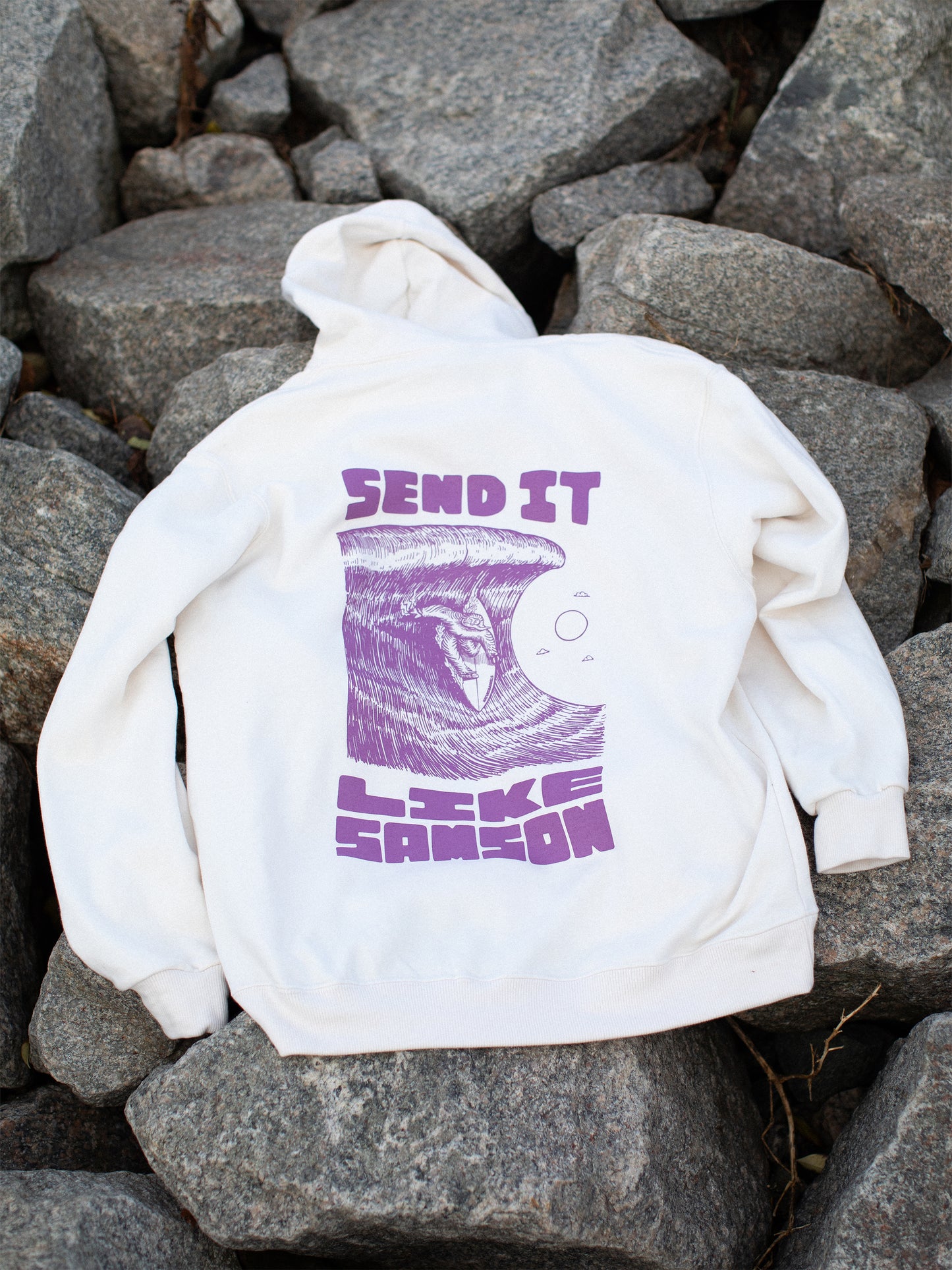 White hoodie with large purple graphic in the back featuring 'SEND IT LIKE SAMSON' text and an illustration of a sasquatch riding a large wave, laid on a rocky surface.