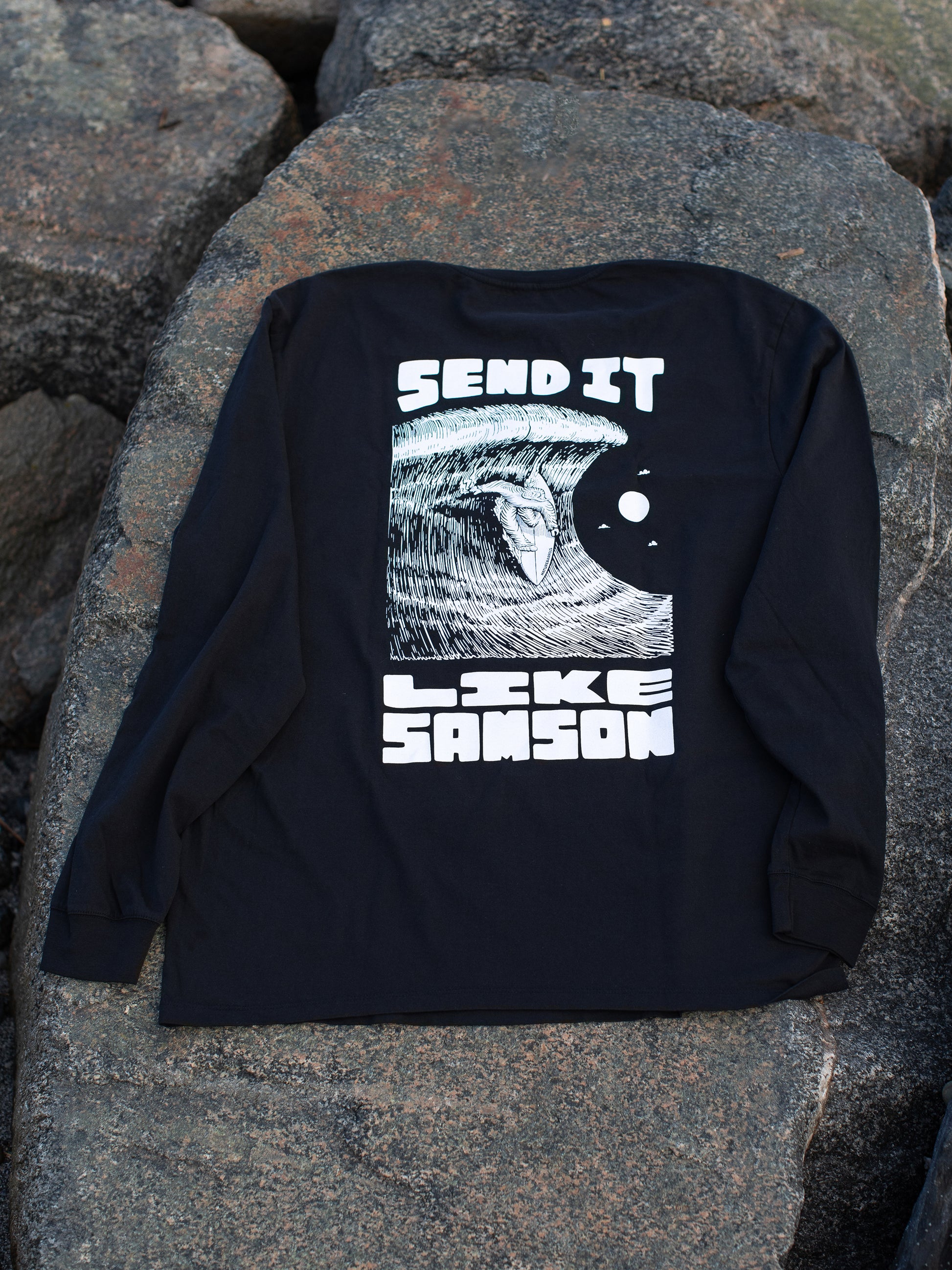 Black long sleeve t-shirt with large white graphic in the back featuring 'SEND IT LIKE SAMSON' text and an illustration of a sasquatch riding a large wave, laid on a rocky surface.