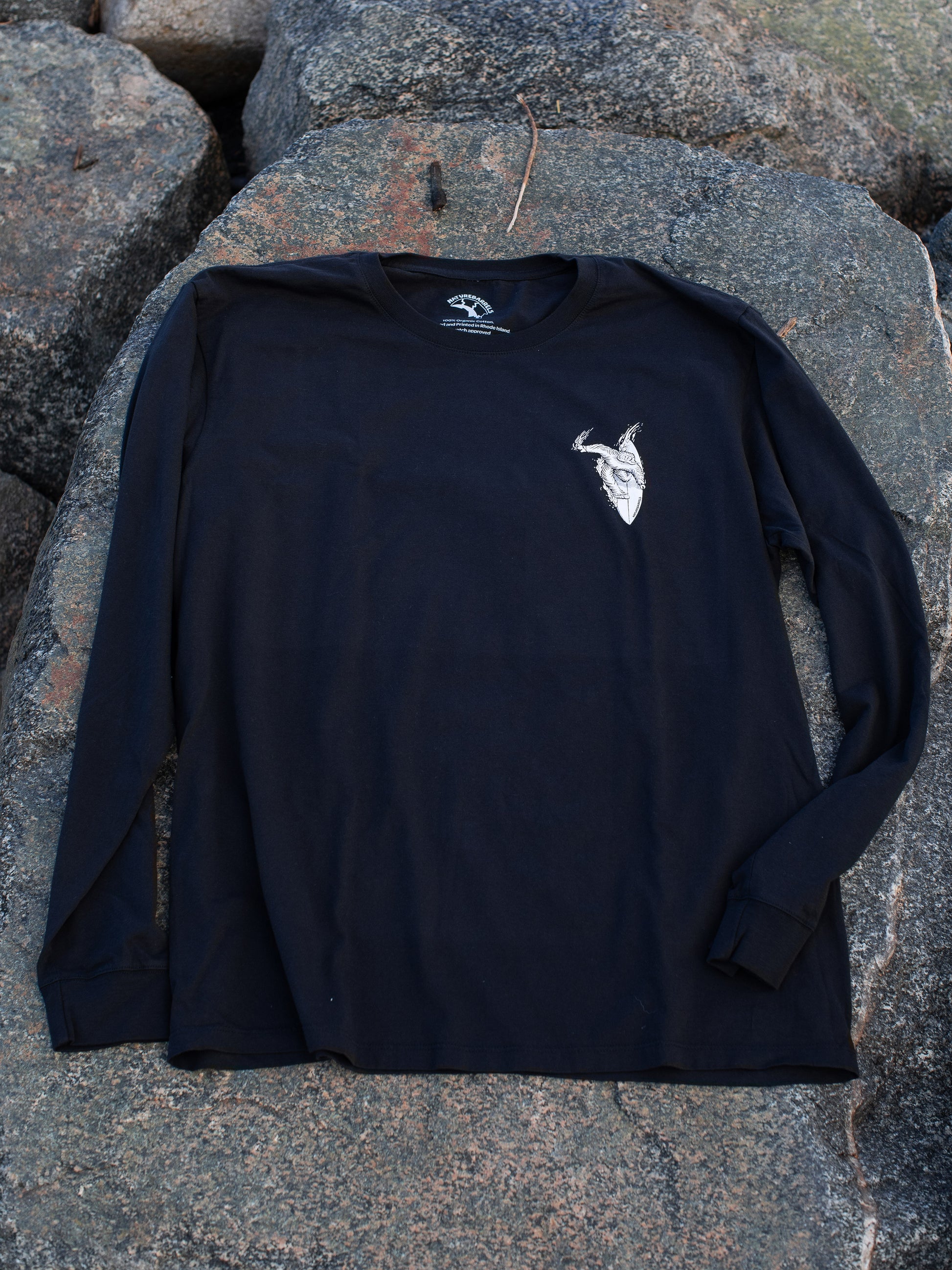 Black long sleeve t-shirt with a small white chest graphic of a surfing sasquatch. Laid on a rocky surface