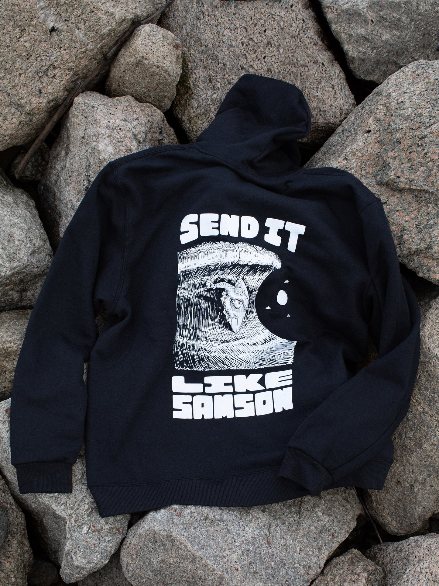 Black hoodie with large white graphic in the back featuring 'SEND IT LIKE SAMSON' text and an illustration of a sasquatch riding a large wave, laid on a rocky surface.