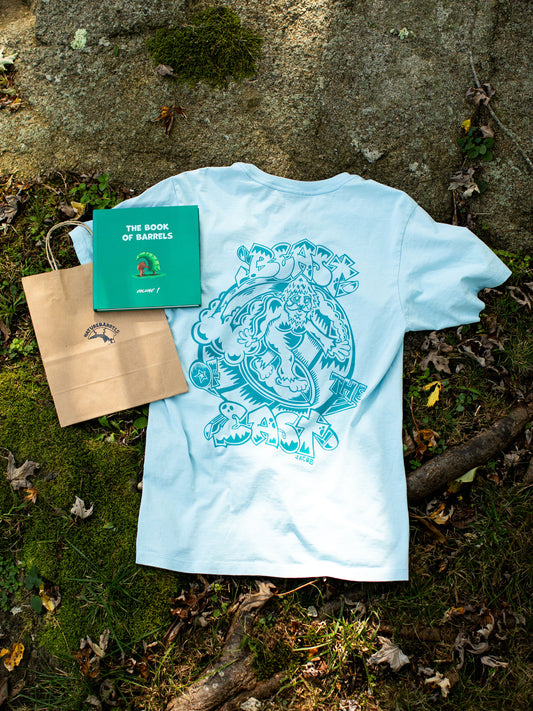 A light blue t-shirt displayed flat on a natural ground of moss and leaves, featuring a large, detailed back print in darker blue that includes a surfing sasquatch riding a wave. Next to the t-shirt is a green hardcover book titled "the book of barrels, volume 1" and a recycled brown paper bag with the brand's logo on it