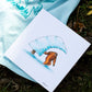 A vibrantly colored illustration from 'The Book of Barrels' showing an orange sasquatch under a snow-covered pine tree placed atop the back of a light blue t-shirt with a large dark blue graphic. The t-shirt and book lie on the natural forest floor.