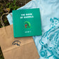 A top view of a hardcover book 'The Book of Barrels, Volume 1' showcasing an illustration of an orange sasquatch under a wave-shaped pine tree. The book is placed on the back of a light blue t-shirt with a thematic, intricate graphic. The book is adjacent to a brown paper bag with the brand's logo, on an outdoor setting.