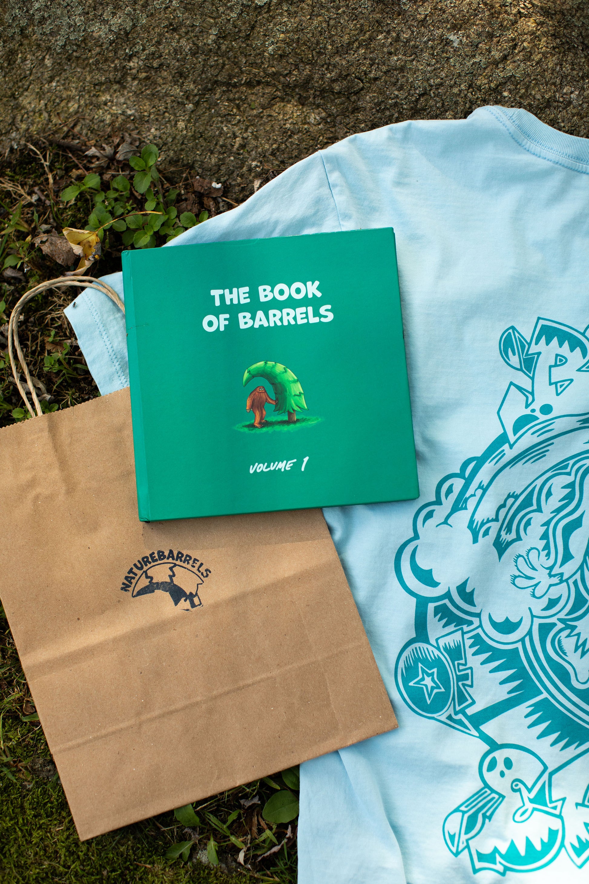 A top view of a hardcover book 'The Book of Barrels, Volume 1' showcasing an illustration of an orange sasquatch under a wave-shaped pine tree. The book is placed on the back of a light blue t-shirt with a thematic, intricate graphic. The book is adjacent to a brown paper bag with the brand's logo, on an outdoor setting.
