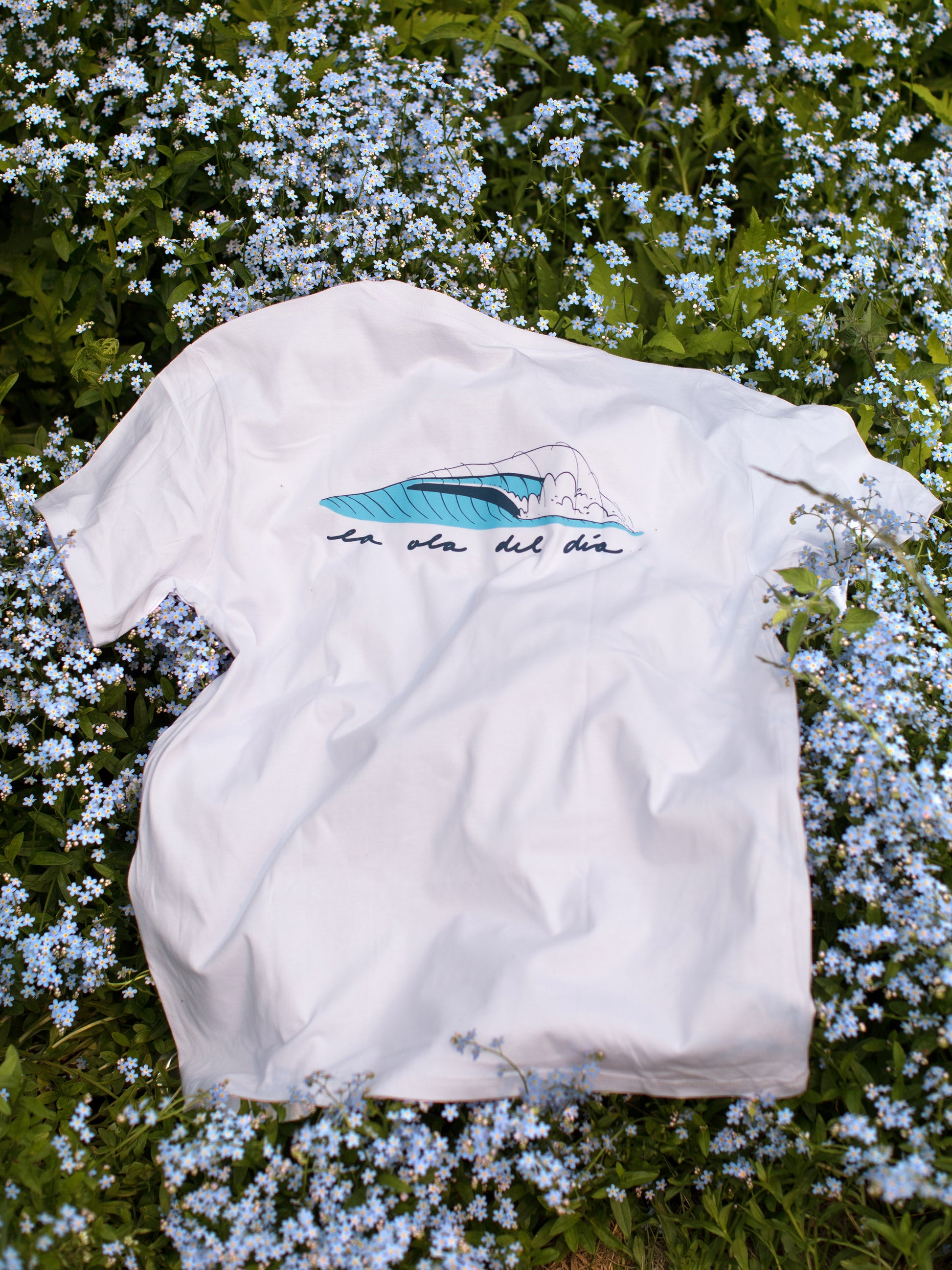 Back view of a white t-shirt with a large blue barrel wave design and text written in Spanish reading "la ola del día" (which means 'the wave of the day'), lying on a bed of small blue flowers.