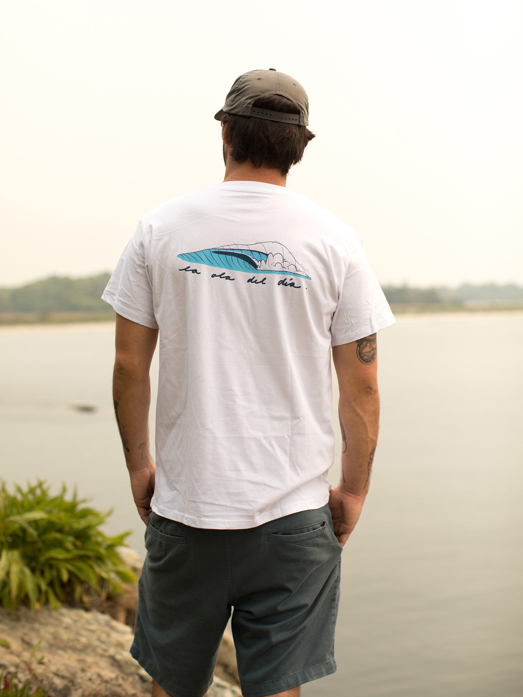 Man with his back towards the camera, wearing a white t-shirt with a blue wave graphic and text 'la ola del día' on the back, standing by the water.