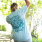 An outdoors photograph of a woman wearing a light blue t-shirt showcasing a large, detailed graphic print in a contrasting darker blue on the back. The print features bold text that reads "Beast of the East" and an intricate design featuring Samson the Legendary Surfing Sasquatch.