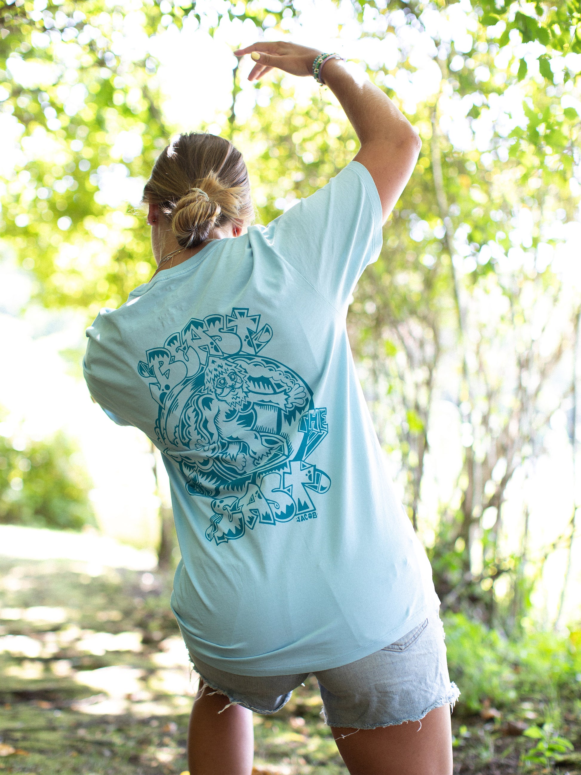 An outdoors photograph of a woman wearing a light blue t-shirt showcasing a large, detailed graphic print in a contrasting darker blue on the back. The print features bold text that reads "Beast of the East" and an intricate design featuring Samson the Legendary Surfing Sasquatch.
