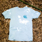 Light blue t-shirt lying flat on a bed of dried bamboo leaves, with a small logo printed on the upper left side on contrasting dark blue. The logo reads "Nature Barrels" in the style of artist Andrew Jacob.   The shirt is centered in the frame, with a hint of sunlight filtering through the foliage above, creating patterns of light and shadow on the fabric and the ground around it.