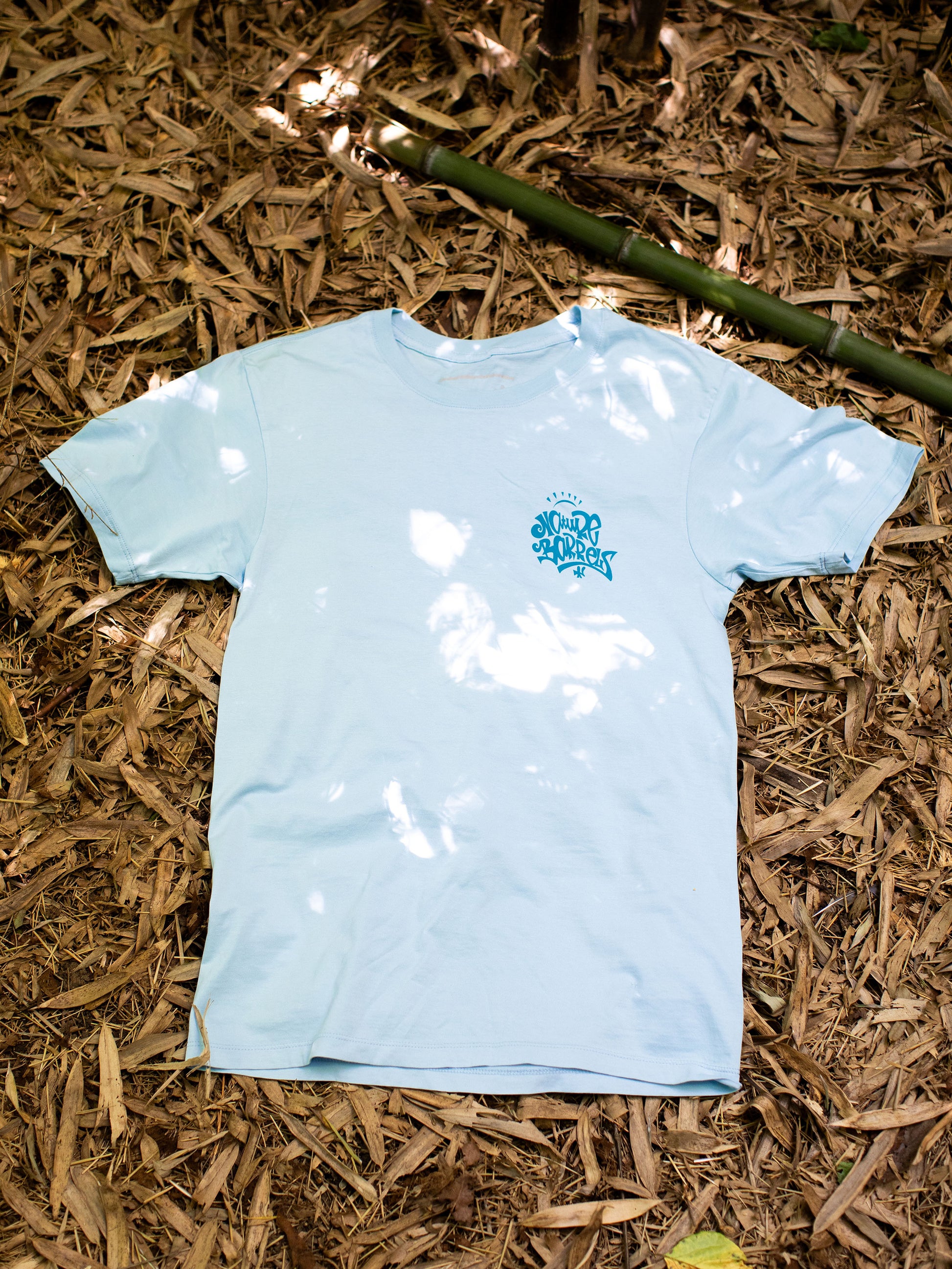 Light blue t-shirt lying flat on a bed of dried bamboo leaves, with a small logo printed on the upper left side on contrasting dark blue. The logo reads "Nature Barrels" in the style of artist Andrew Jacob.   The shirt is centered in the frame, with a hint of sunlight filtering through the foliage above, creating patterns of light and shadow on the fabric and the ground around it.