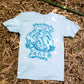 Light blue t-shirt displayed flat on a background of dry bamboo leaves, showcasing a large, detailed graphic print in a contrasting darker blue on the back. The print features bold text that reads "Beast of the East" and an intricate design featuring Samson the Legendary Surfing Sasquatch.