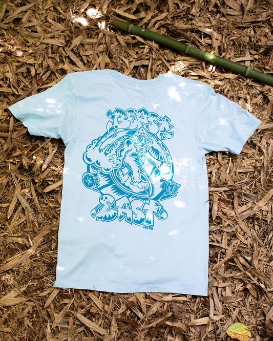 Light blue t-shirt displayed flat on a background of dry bamboo leaves, showcasing a large, detailed graphic print in a contrasting darker blue on the back. The print features bold text that reads "Beast of the East" and an intricate design featuring Samson the Legendary Surfing Sasquatch.