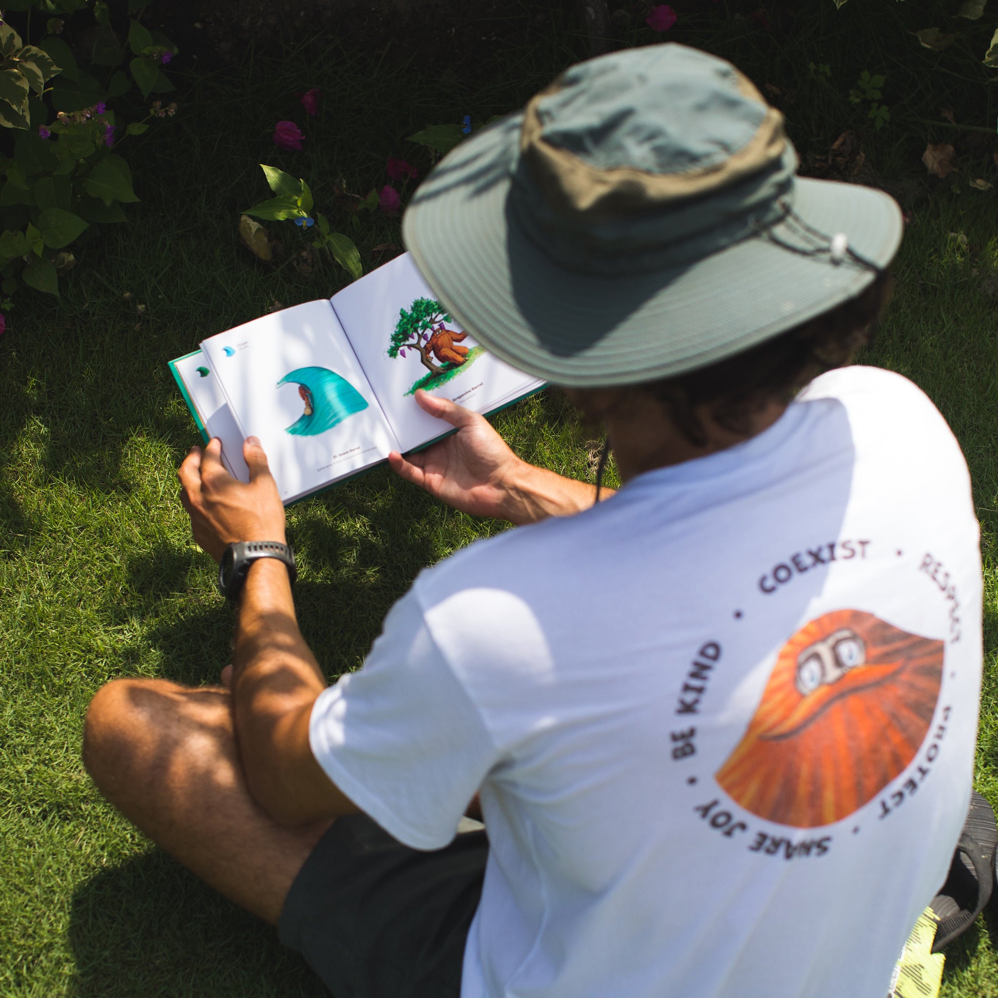A person in a white t-shirt and a bucket hat is reading an "The Book of Barrels" in the grass, with illustrations visible on the pages as they enjoy a sunny day outdoors.