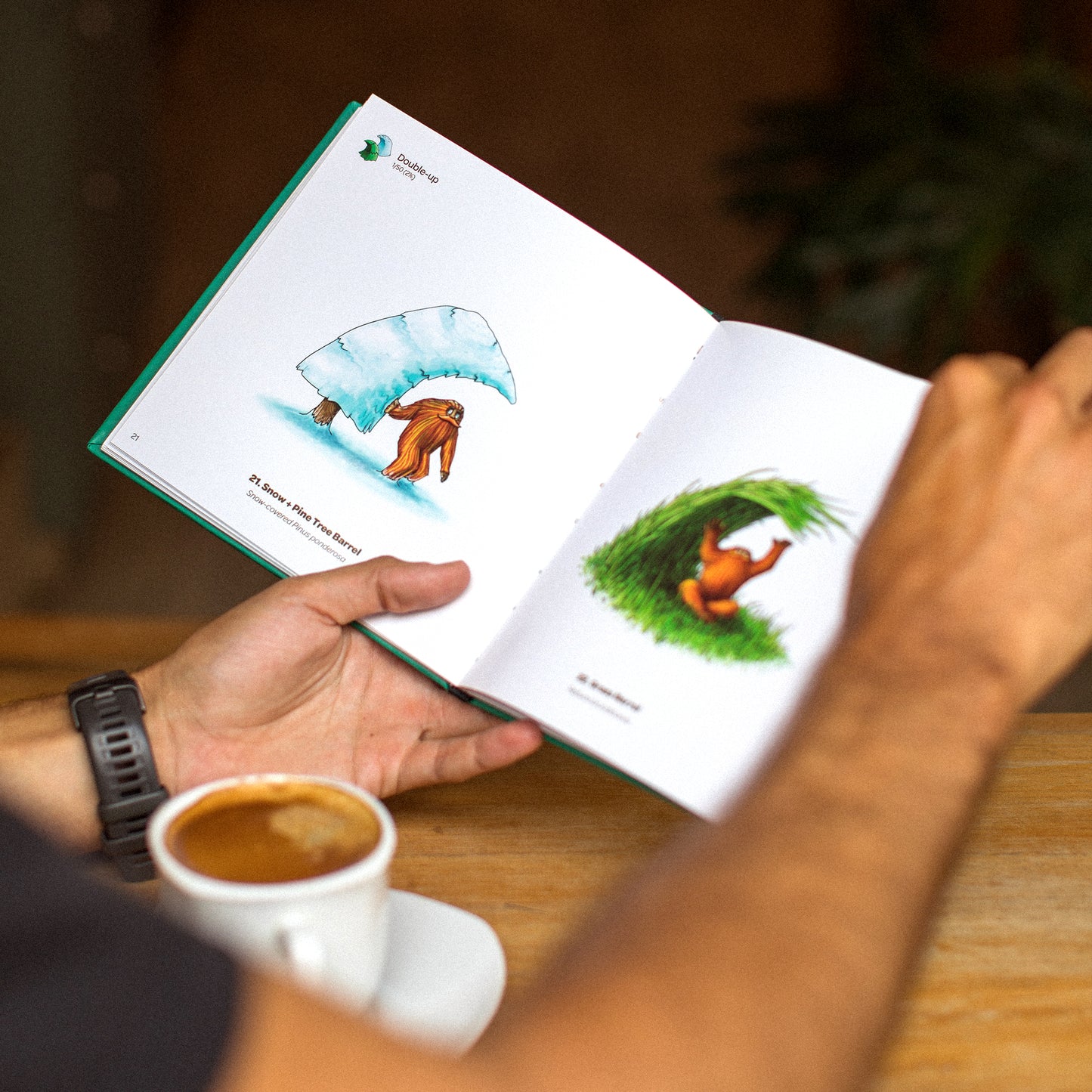 An open hardcover book on a wooden table showing two pages, with illustrations of a figure under a snow-covered pine tree wave on the left and a figure under tall grass in the shape of a wav,. There is a cup of espresso on the side.