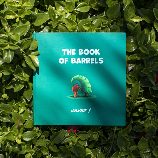 A hardcover book titled 'The Book of Barrels, Volume 1' is placed on a lush bed of green leaves, with a colorful illustration of a Sasquatch standing under a wave-shaped pine tree  on the cover.