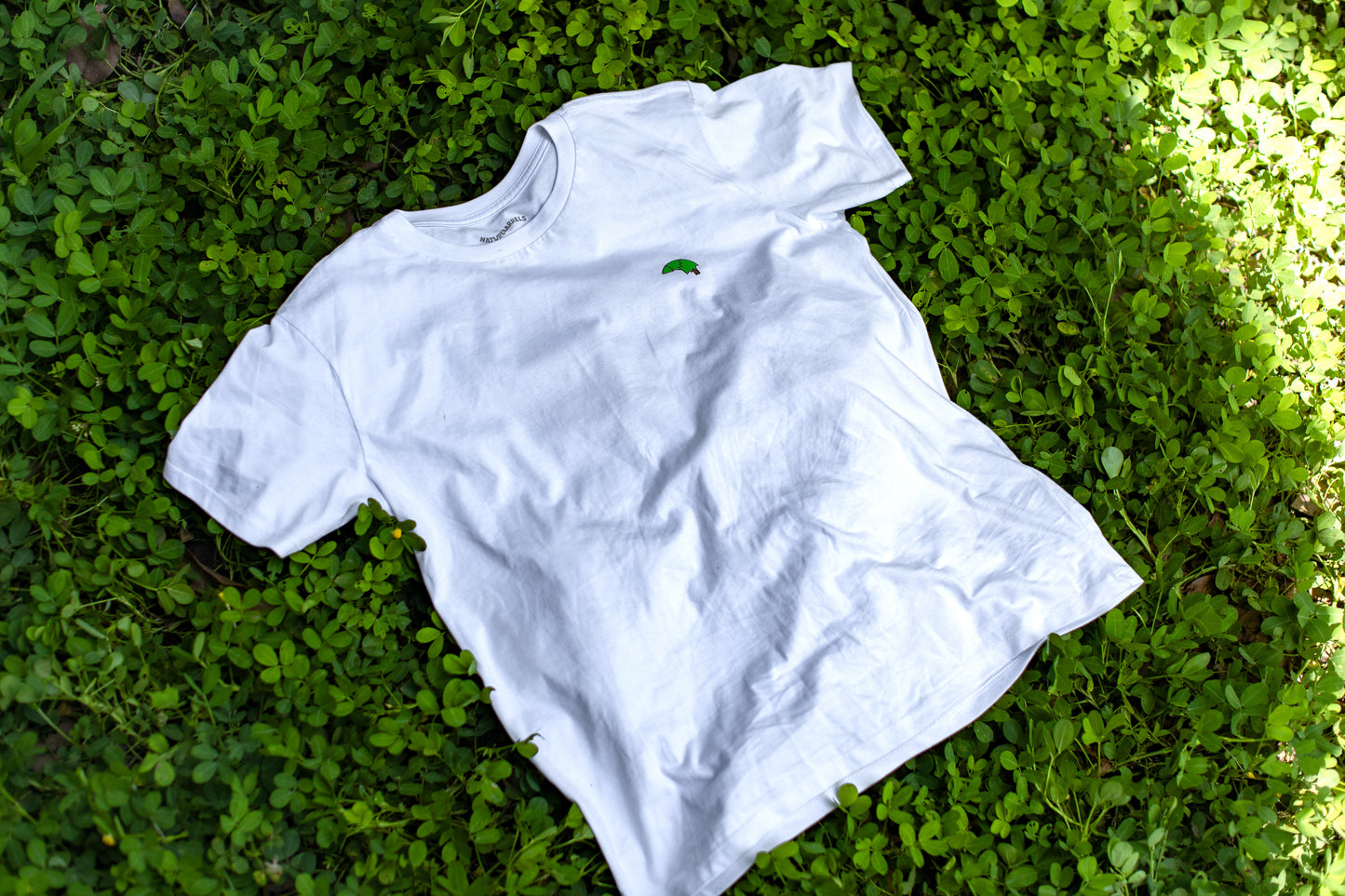 A plain white t-shirt with a small 'Naturebarrels' chest logo lying on a bed of green clover leaves