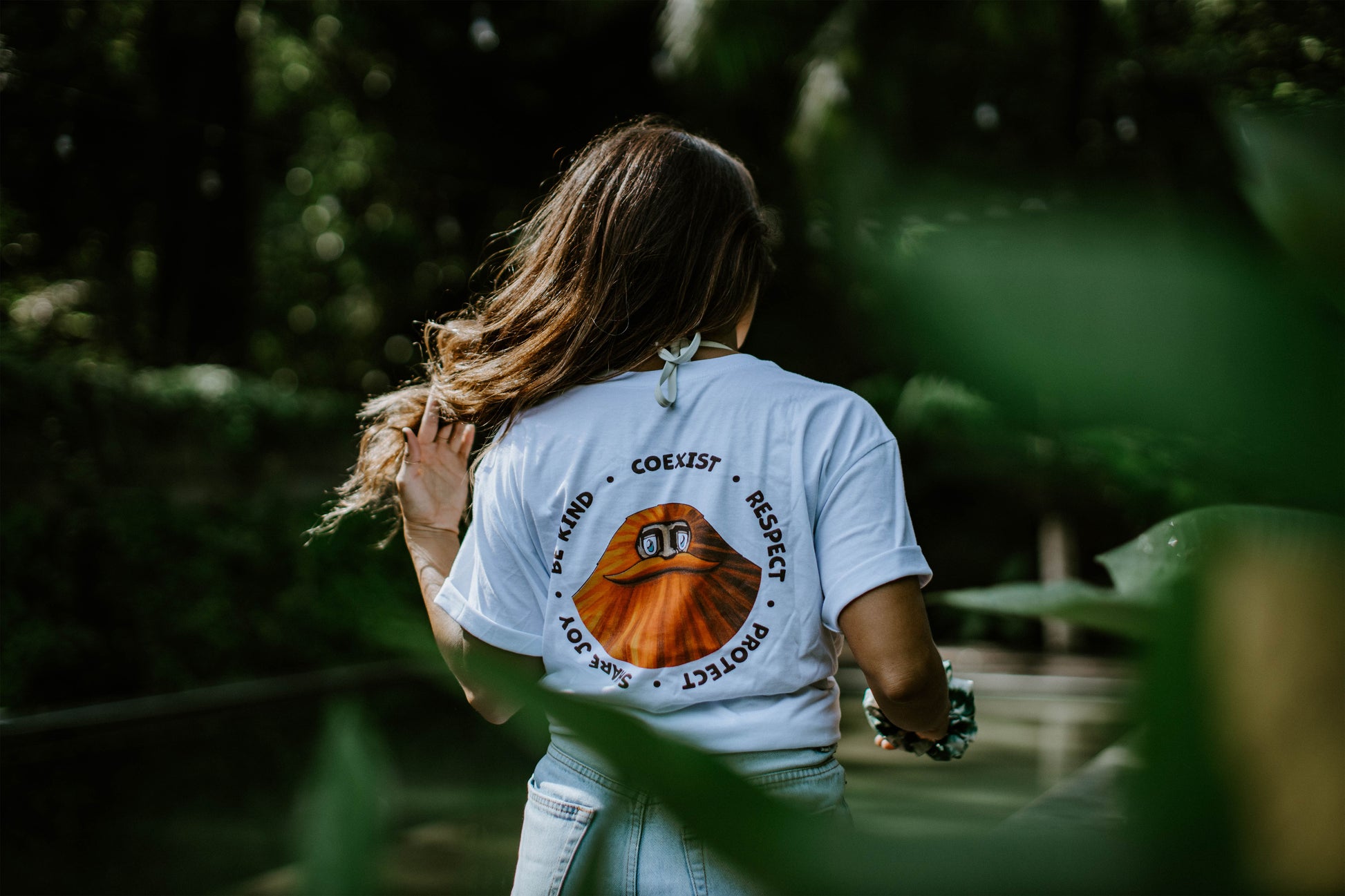 A person statheding with his back towards the camera,   A woman with their back towards the camera wearing a white t-shirt with a print of an orange sasquatch with a great moustache and text in a circular formation around it that reads 'Share Joy, Be Kind, Coexist, Respect, Protect'